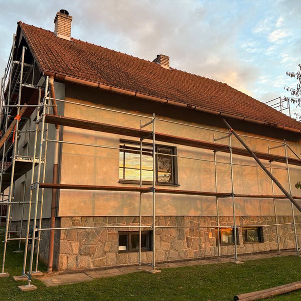Fasádní lešení na rodinném domě s červenou střešní krytinou, večerní světlo